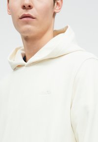 Jovem vestindo um hoodie de cor creme com um pequeno logótipo em relevo da ellesse no peito, contra um fundo simples.