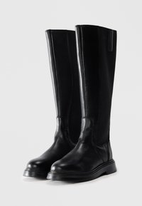 Une paire de bottes en cuir noir hautes avec des orteils ronds et des semelles en caoutchouc plates, se tenant debout sur un fond neutre.