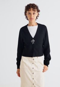 Cardigan tricotat negru cu decolteu în V și mâneci lungi, dotat cu un laț în față. Purtați-l peste un top alb, asortat cu o fustă bej cu nasturi.