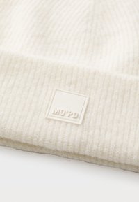 Krēmveida adīta cepure ar ribotu tekstūru, kurai ir kvadrātveida balta uzlīme ar izsistiem burtiem "MO'PD". Audums izskatās mīksts un biezs.