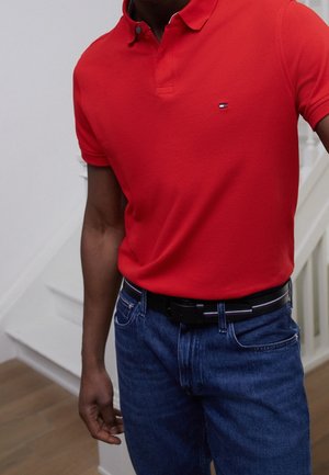 Tee-shirt polo rouge avec un col, arborant un petit emblème sur la poitrine. Porté avec un jean bleu et une ceinture noire. Design ajusté et texture lisse.
