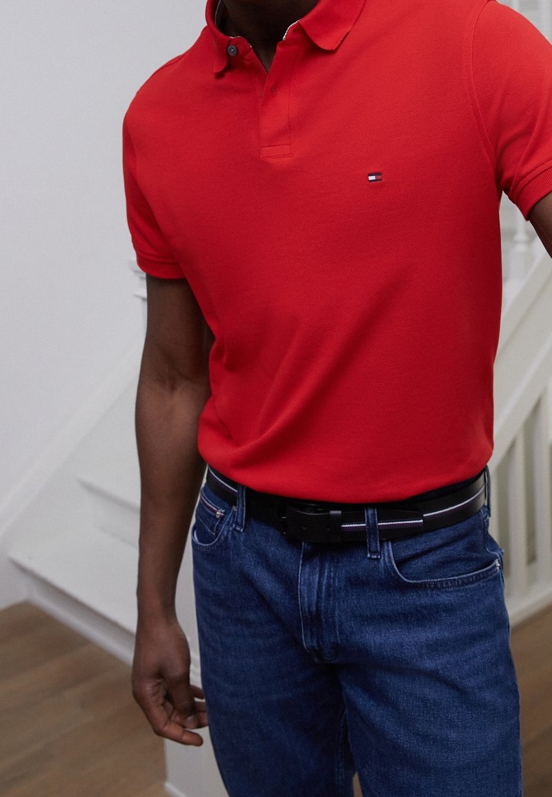 Tee-shirt polo rouge avec un col, arborant un petit emblème sur la poitrine. Porté avec un jean bleu et une ceinture noire. Design ajusté et texture lisse.