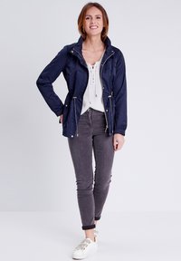 Giacca impermeabile blu navy con zip frontale, vita regolabile con cordino e tasche laterali, abbinata a jeans skinny grigi e sneakers bianche.