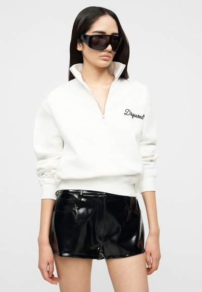 Femme portant de grandes lunettes de soleil noires, un sweat-shirt blanc à demi-fermeture éclair avec logo, et un short en cuir noir brillant, debout devant un fond uni.