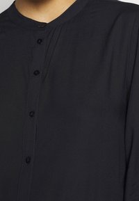 Svart skjorta med mandarin-krage, tyg med mjuk textur och sex svarta knappar nedanför.