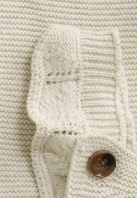 Gros plan d'un tissu tricoté de couleur crème avec des motifs en maille câble et en côtes, mettant en avant un grand bouton marron à quatre trous.