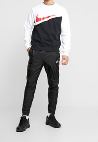 Svart och vit Nike-tröja med en stor röd logotyp, i kombination med glänsande svarta träningsbyxor och svarta sneakers med vita accenter.