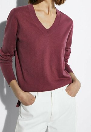 Personne portant un pull bordeaux à manches longues et col en V, rentré dans un pantalon blanc taille haute, les mains dans les poches.