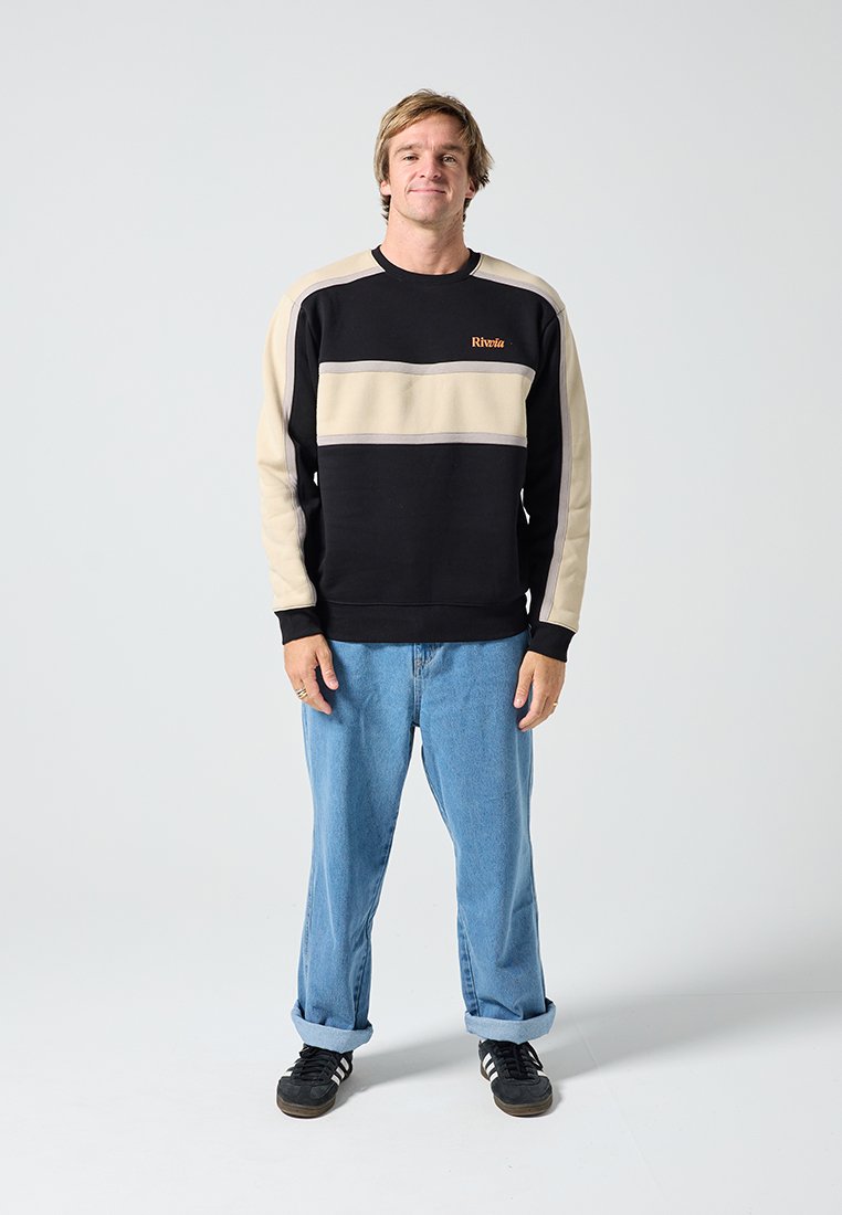 Φούτερ σε μαύρο και μπεζ με μπλοκ χρωμάτων, μακριά μανίκια και ριμπ μανσέτες, με πορτοκαλί λογότυπο. Φορεμένο με ανοιχτό μπλε τζιν και μαύρα παπούτσια.