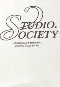 Weißer Stoff mit dunkel gedrucktem Text: "STUDIO. SOCIETY" in großer Schrift und "SCHAFFE EIN LEBEN, FÜR DAS DU NICHT WARTEN KANNST, UM DARAUF WACHZU WERDEN." in kleinerer Schrift.