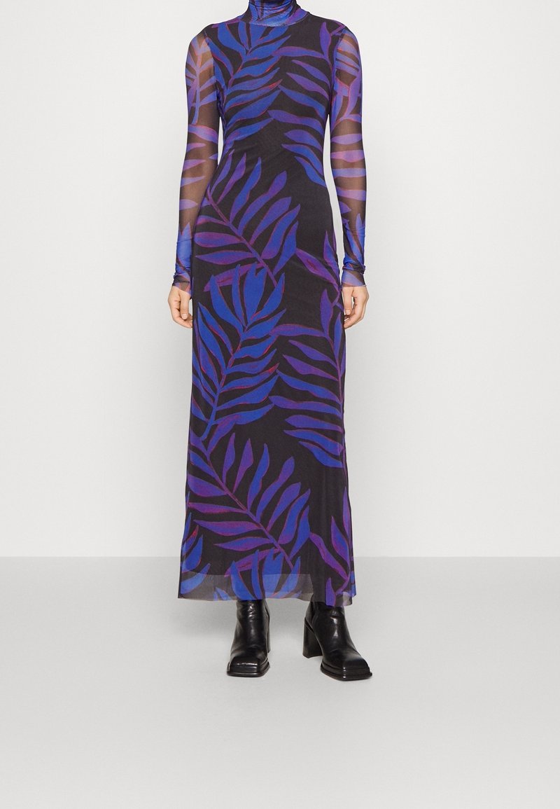 Femme portant une longue robe noire transparente avec des motifs de feuilles bleues et violettes et des bottines noires, debout devant un fond uni.