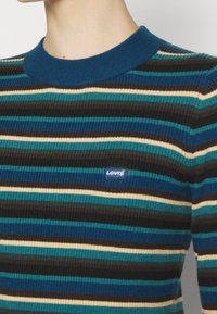 Gros plan d’un pull en maille côtelée avec des rayures horizontales en bleu, sarcelle, marron et crème, avec une étiquette logo Levi’s bleue sur la poitrine.