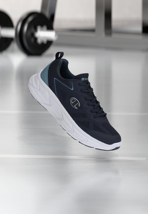 Sneaker atletico nero e verde acqua con suola bianca spessa, logo del marchio sul lato, sospeso sopra un pavimento da palestra riflettente con pesi sfocati sullo sfondo.