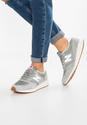 Person trägt graue und weiße New Balance 420 Sneakers mit hochgekrempelten blauen Jeans auf einem hellen Boden, ein Fuß leicht angehoben.