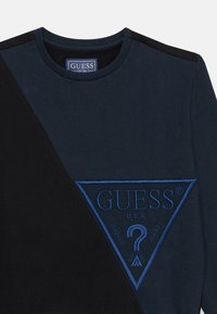 Svart og marineblå genser med rund hals, med stort blått brodert trekantlogo som har "GUESS" og et spørsmålstegn som detalj.