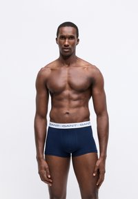 Uomo muscoloso in piedi contro uno sfondo bianco, indossa boxer in blu navy GANT con elastico bianco e logo.
