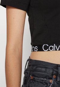 Musta lyhythihainen crop-top, jossa on valkoinen "Calvin Klein" -logo vyötäröllä. Yhdistetty tummansinisiin korkeavyötäröisiin farkkuihin.