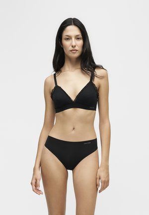 Frau mit langen dunklen Haaren, die einen schwarzen Bralette und passende schwarze Unterwäsche trägt, steht vor einem schlichten hellen Hintergrund.