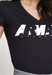 Černé V-šité tričko s velkým bílým nápisem "ARMANI EXCHANGE", které nosí osoba s dlouhými tmavými vlasy a stříbrným přívěskem na náhrdelníku.