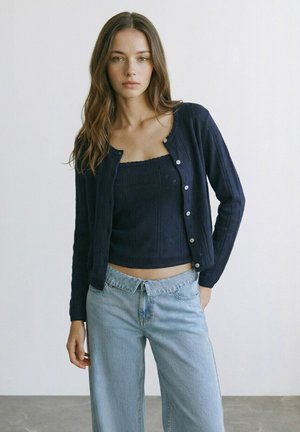 Jonge vrouw met lang bruin haar, gekleed in een marineblauw vest met knopen over een bijpassende top en lichtblauwe spijkerbroek, staand tegen een witte muur.