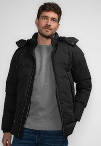 Giacca a vento nera con cappuccio rimovibile, zip frontale e polsini elasticizzati. Indossata sopra un maglione grigio e abbinata a jeans blu.