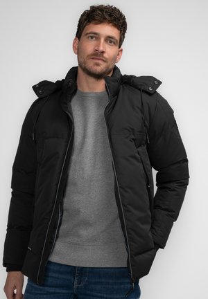 Schwarze Puffjacke mit abnehmbarer Kapuze, Reißverschluss vorne und elastischen Bündchen. Getragen über einem grauen Pullover und kombiniert mit blauen Jeans.
