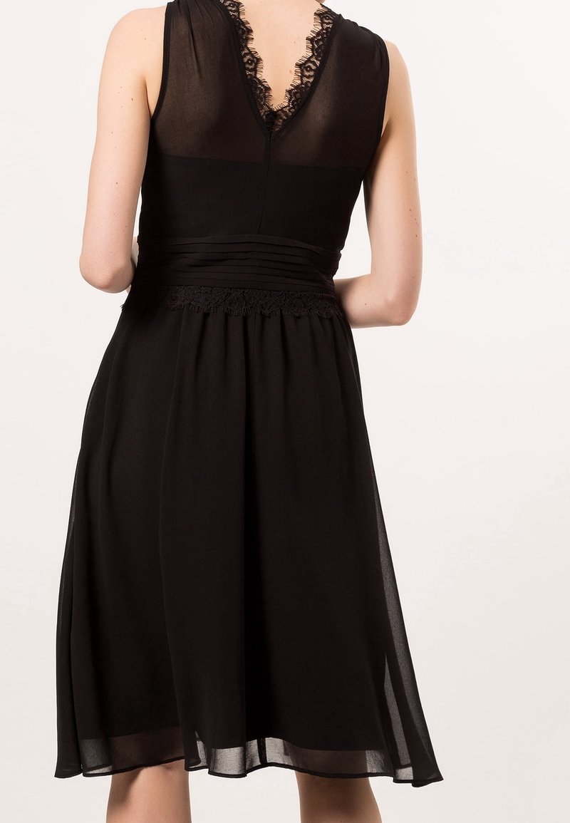 Robe noire sans manches avec un dos en V en dentelle transparente, taille ajustée avec des plis et détails en dentelle, et une jupe fluide jusqu'au genou.