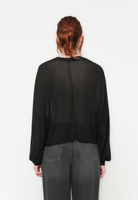 Blouse noire transparente à manches longues et ourlet arrondi, avec un détail de couture subtil à l'arrière. Associée à un jean gris taille haute.