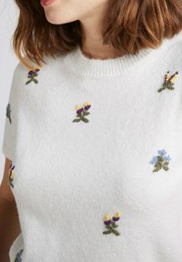 Pull blanc à manches courtes, décoré de broderies florales multicolores en jaune, violet et bleu. Texture douce, encolure côtelée.