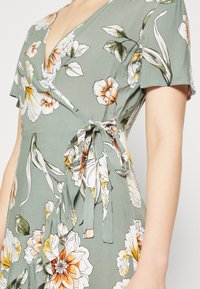 Kwiatowa sukienka wrap w zielonym kolorze, z białymi i pomarańczowymi kwiatami, głębokim dekoltem w serek, krótkimi rękawami oraz detalem związanym z boku.