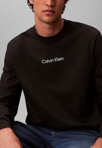 Hombre con sudadera negra de Calvin Klein y vaqueros azules, sentado frente a un fondo gris liso.
