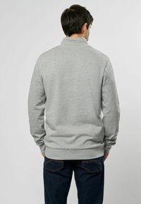 Lys grå hettegenser med høy krage, myk teksturert stoff, ribbestrikkede mansjetter og kant. Bæreres over mørke jeans, sett fra baksiden.