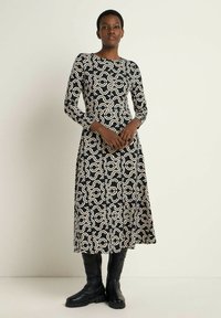 Langärmliges schwarzes Kleid mit cremefarbenem abstraktem Muster, A-Linie Silhouette, Rundhalsausschnitt, kombiniert mit knielangen schwarzen Stiefeln.
