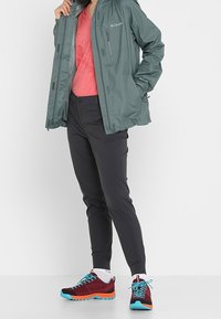 Osoba v zelené bundě Columbia, červeném tričku, černých kalhotách a červenobílých teniskách stojí na jednolitém pozadí.