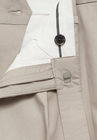 Gros plan d’un pantalon beige avec une fermeture éclair visible, bouton, fermeture par crochet métallique et une partie de la doublure intérieure blanche avec un détail de bande noire.