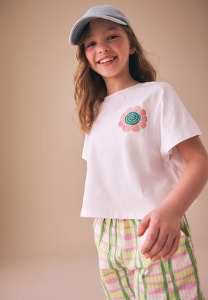 Fille souriante portant une casquette grise, un t-shirt blanc avec un motif de fleur rose et verte, et un pantalon rayé pastel rose, vert et jaune.