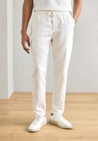 Hvide linned træksnore bukser med en afslappet pasform og sidelommer, parret med hvide sneakers med brune detaljer.