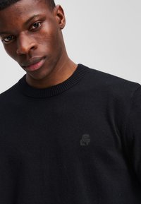 Jersey de punto negro con cuello redondo y un logotipo sutil en el pecho. Presenta una tela texturizada y puños acanalados.