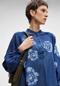 Πουκάμισο τζιν σε μπλε χρώμα με λευκό λουλουδάτο μοτίβο, διαθέτοντας γιακά, κουμπιά και φουσκωτά μανίκια. Κάθετες ραφές και υφασμάτινη υφή.