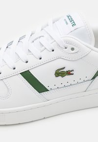 White leather sneaker with green accents, flat laces, perforated details, and embroidered crocodile logo on the side. Simple, low-profile design.