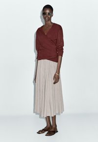 Maroon wrap sweater with long sleeves, paired with a beige pleated skirt. The outfit is complemented by brown slide sandals and dark sunglasses.
