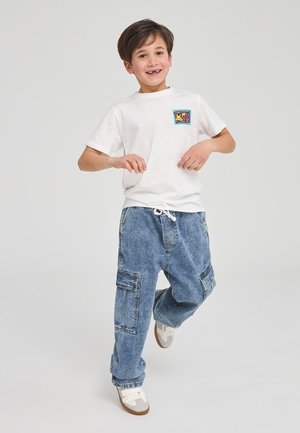 T-shirt bianco a maniche corte con una grafica colorata sul petto, abbinato a pantaloni cargo in denim blu larghi con tasche laterali.