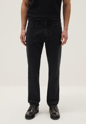 Una persona che indossa jeans neri a gamba dritta, camicia nera e scarpe nere con lacci, in piedi davanti a uno sfondo chiaro e semplice.