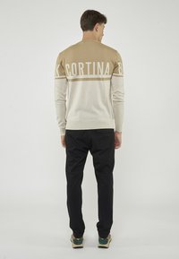 Maglione lavorato a maglia beige e crema con "CORTINA" in lettere grasse sulla schiena, abbinato a pantaloni neri e scarpe multicolori.