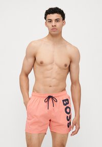 Koralne kopalne hlače iz lahkega materiala, z vrvico v pasu in črnim logotipom "BOSS" na levi strani.
