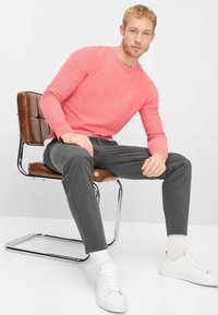 Lichtroze gebreide trui, taps toelopende grijze broek, witte sneakers, zittend op een bruine kussenstoel met chromen frame. Gladde texturen en een aansluitend ontwerp.