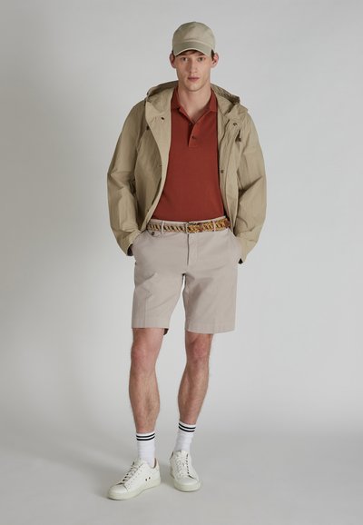 Giacca leggera beige, polo color ruggine, pantaloni corti beige chiaro, cintura a fantasia, sneakers bianche con calzini a strisce nere, sfondo neutro.