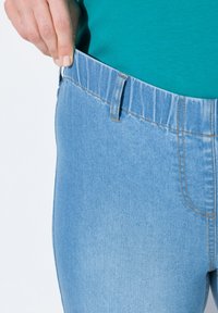 Světlemodré strečové džíny s elastickým pasem, s jemně vybledlou texturou a detaily šití. Ruka držící pas je viditelná.
