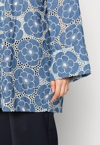 Top de mezclilla con un patrón floral azul, que presenta flores bordadas en blanco y una textura similar a encaje. Mangas acampanadas y un corte holgado.