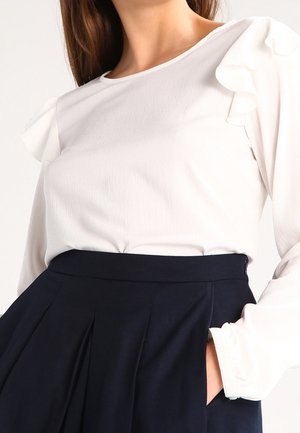 Femme portant un chemisier blanc à manches longues avec des détails froncés aux épaules et une jupe plissée noire taille haute, les mains dans les poches.